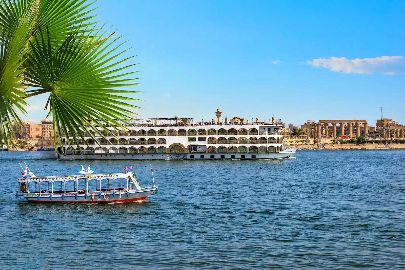A large Nile cruise ship on the river at Luxor, framed by a palm frond, Egypt
